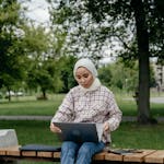 A young Muslim woman in a hijab sitting on a park bench working on a laptop outdoors.