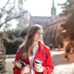 Smiling woman in red coat with coffee and books, enjoying leisure time outdoors on a university campus.